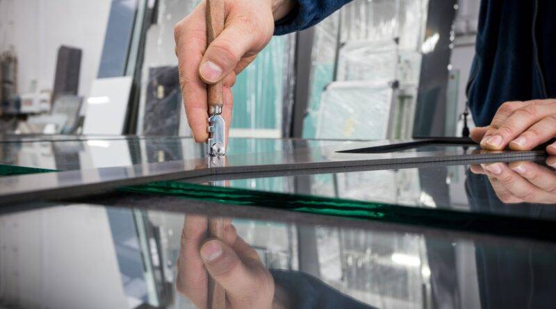 Ein Fensterbauer in Wuppertal schneidet präzise Glas mit einem speziellen Werkzeug. Der Handwerker trägt eine blaue Arbeitsjacke und arbeitet sorgfältig an einer Glasplatte in einer Werkstatt. Im Hintergrund sind weitere Glasplatten und Werkstattutensilien zu sehen.
