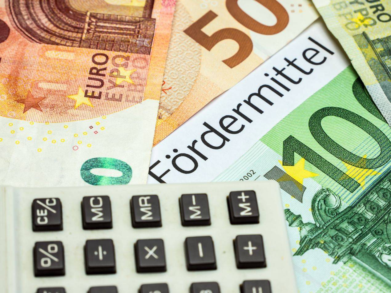 Euro-Geldscheine, ein Taschenrechner und das Wort „Fördermittel“ verdeutlichen finanzielle Unterstützungsmöglichkeiten bei der Baufinanzierung in Mönchengladbach.